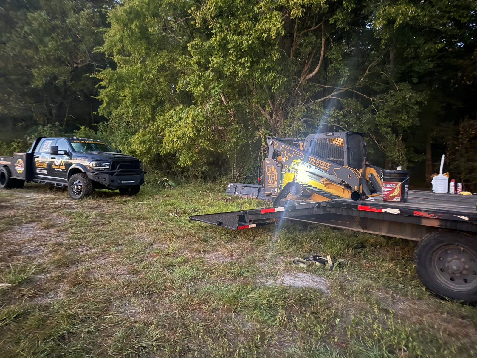 Tracked skid steer loader on flatbed trailer beside custom work truck near wooded clearing site