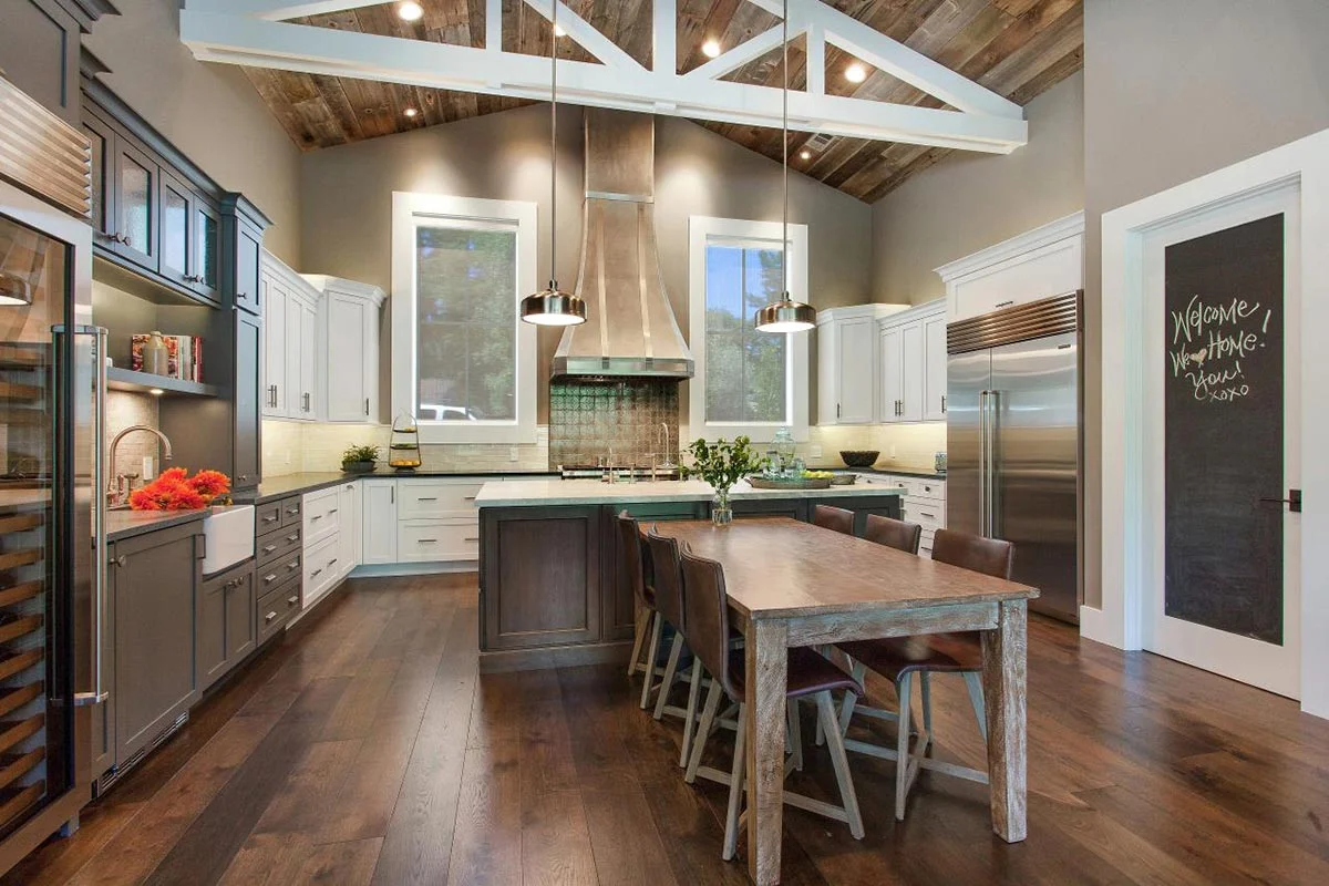 Open kitchen with white shaker cabinets, gray lower cabinets, wood farmhouse dining table, stainless hood, and reclaimed wood vaulted ceiling