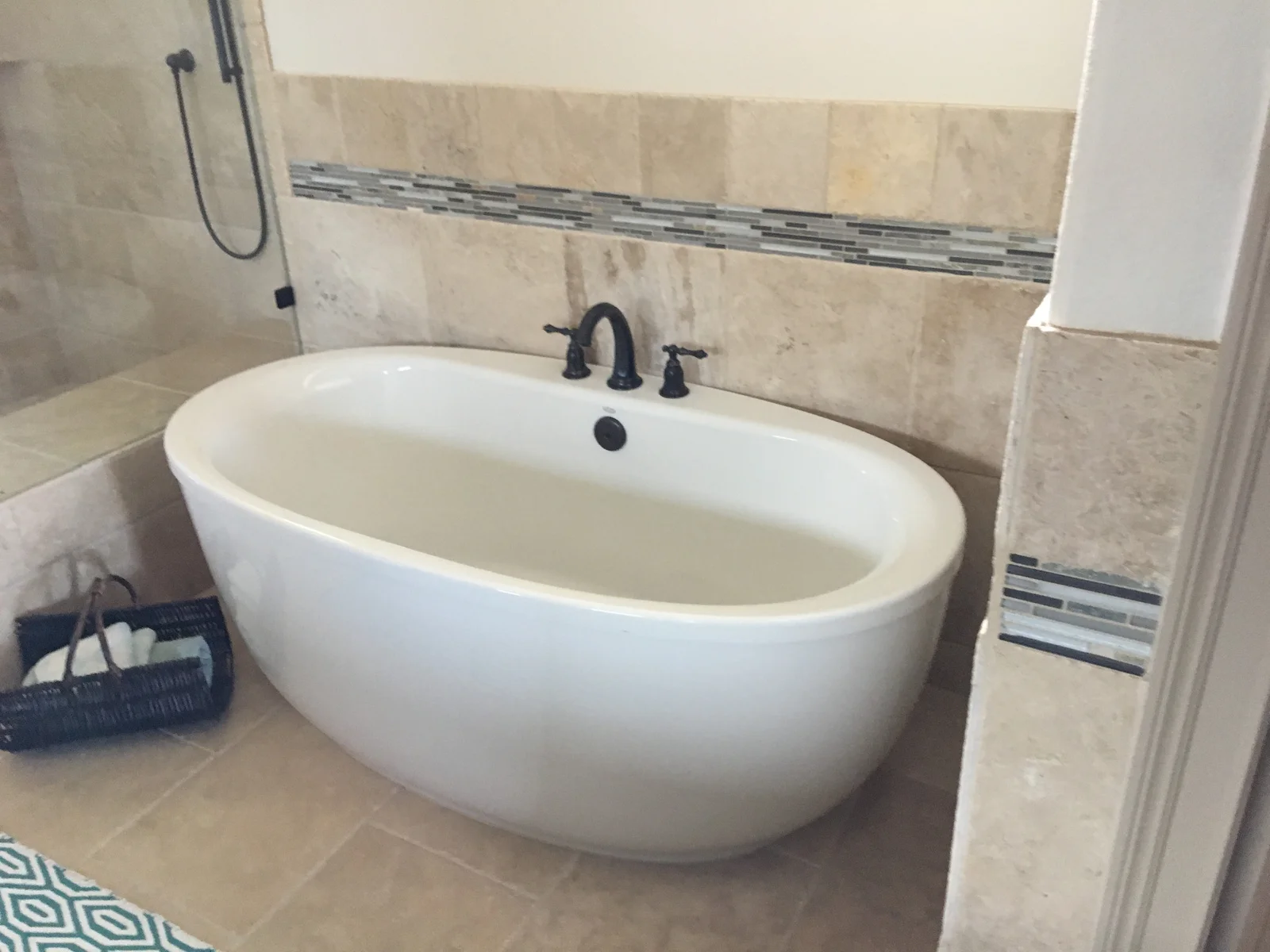 Freestanding white oval tub with bronze faucet against beige tile wall with glass mosaic accent strip