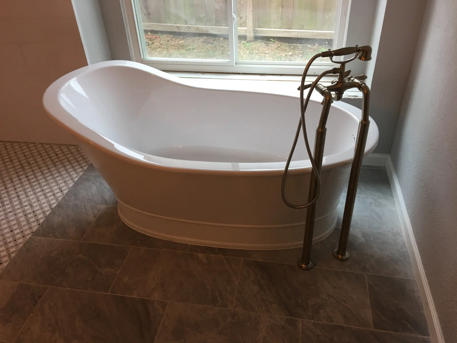White freestanding slipper tub with brushed nickel floor-mount tub filler and handheld shower on gray slate-look tile floor