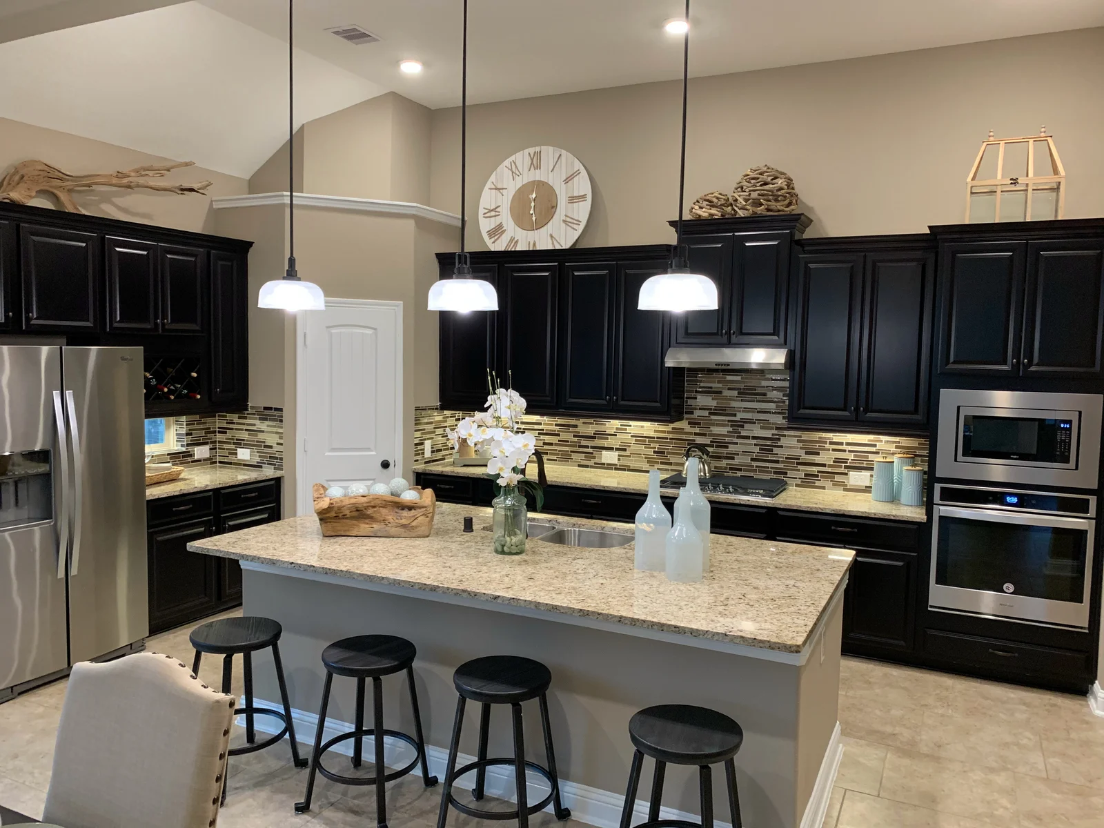 Kitchen with dark raised-panel cabinets, beige granite island, mosaic glass backsplash, and white pendant lights