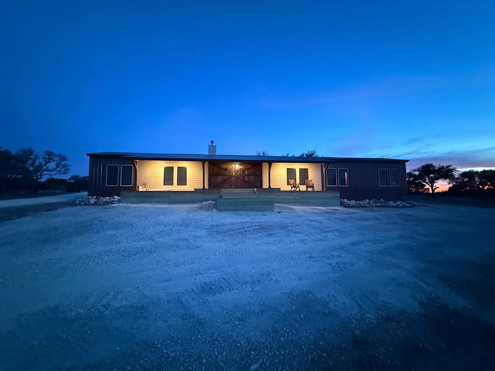 Dusk view of single-story ranch home with dark metal roof, board-and-batten siding, cream lap siding porch, barn-style door, and wood porch steps
