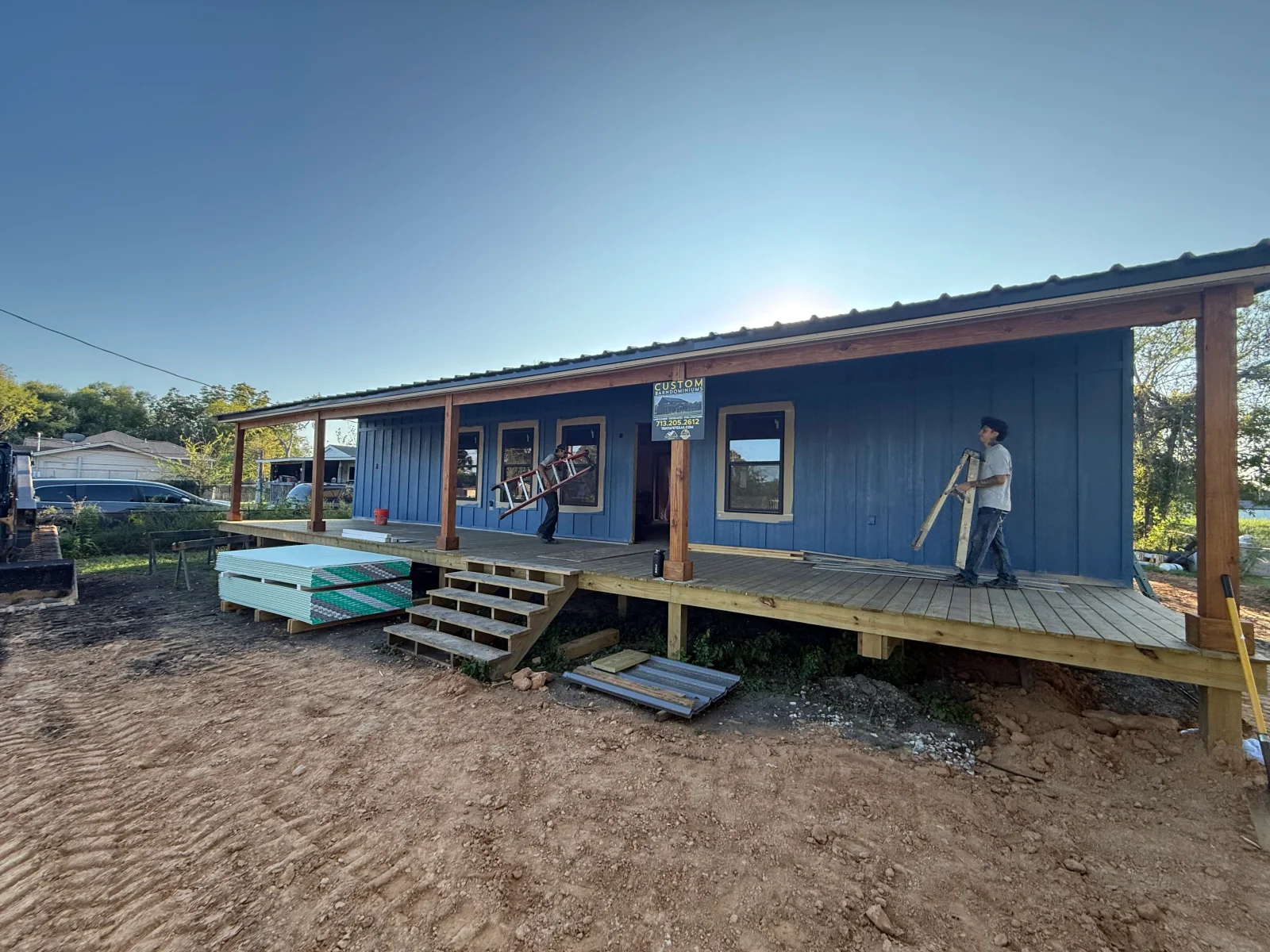 Blue board-and-batten barndominium under construction with timber porch posts, wood deck, and metal roofing