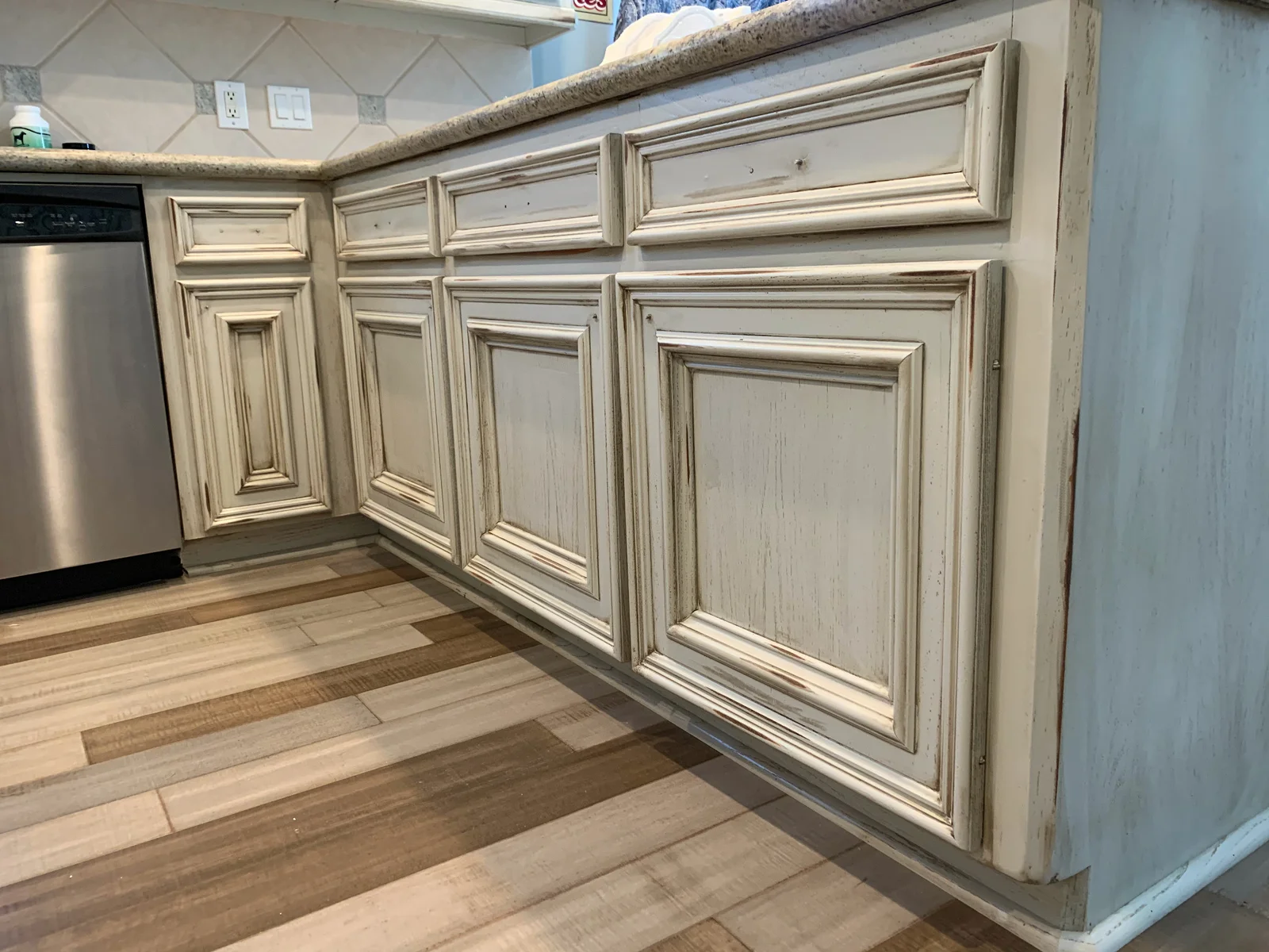 Distressed cream raised-panel kitchen base cabinets with glazed edges, granite countertop, and wood-look plank flooring