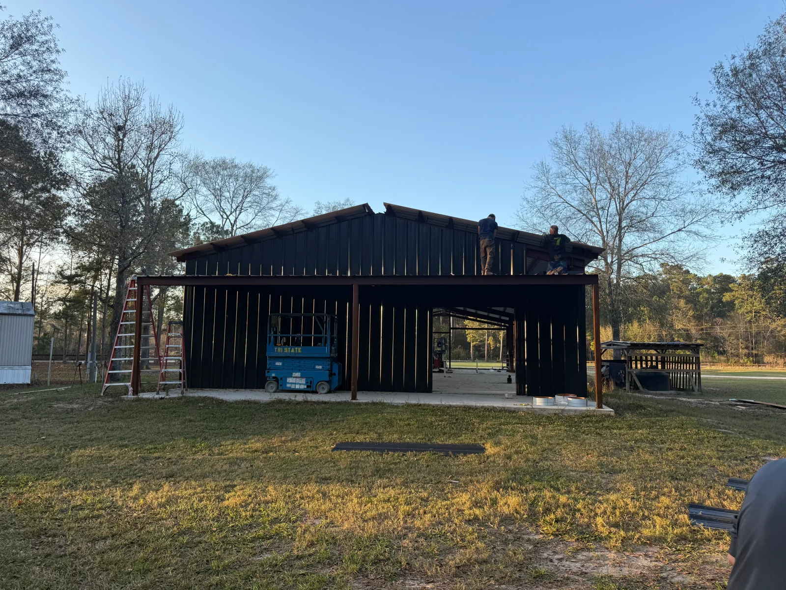 Dark vertical metal siding and gabled steel roof on a pole barn under construction, with a scissor lift and ladders on a concrete slab