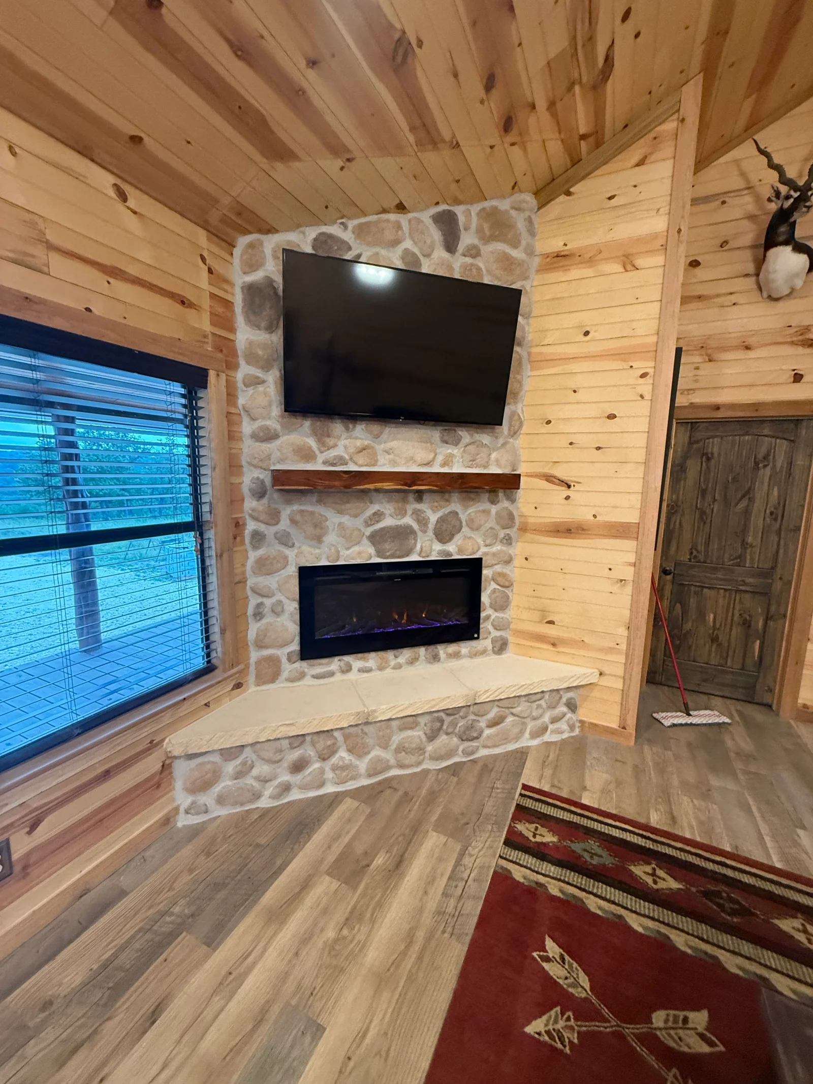 Corner river stone fireplace with live-edge wood mantel, electric insert, and knotty pine paneled walls