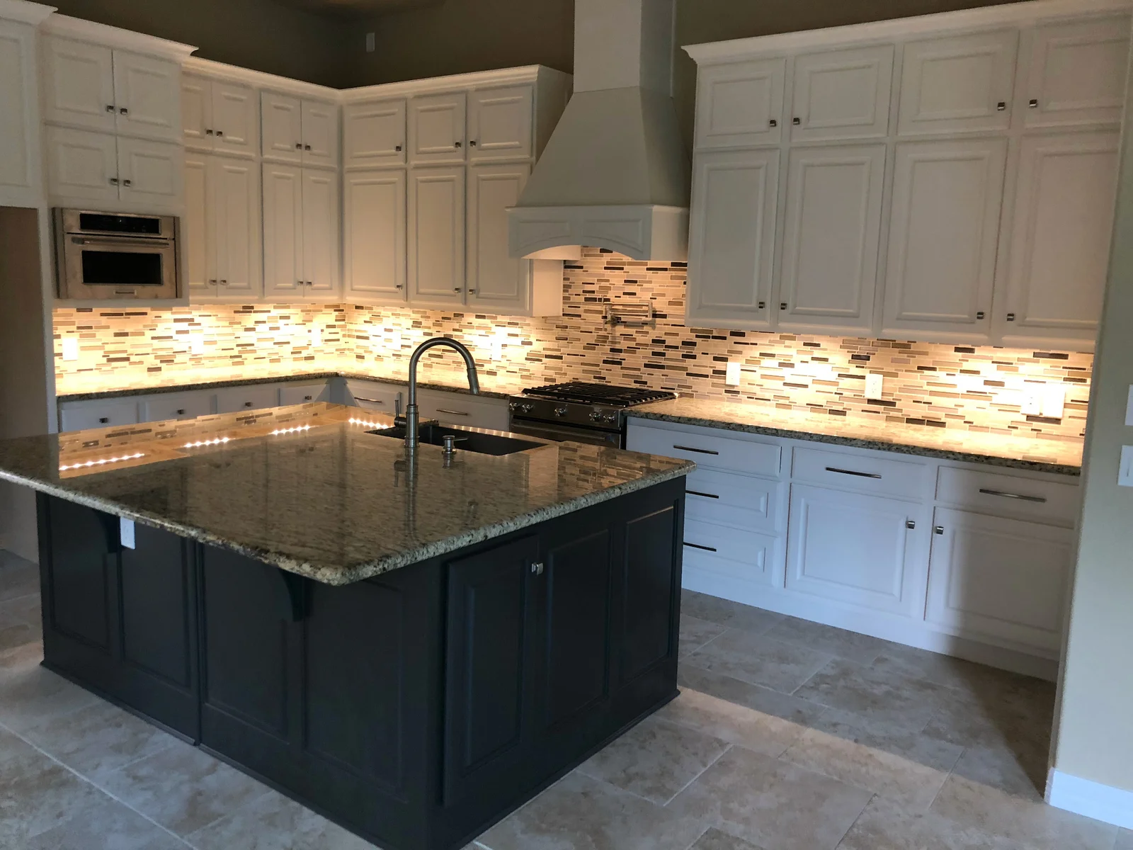 Kitchen with white raised-panel cabinets, dark island, granite countertops, mosaic tile backsplash, and white range hood